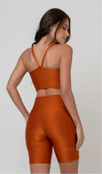 Uma modelo feminina posando de costas contra um fundo cinza neutro, vestindo um conjunto fitness feminino laranja, com proteção solar. O top fitness de academia tem alças finas. A peça de baixo é um short de ciclismo (bermuda fitness feminina) de cintura alta também na cor laranja, com um bolso lateral visível onde a modelo está com a mão direita inserida. Ela olha para o lado. 