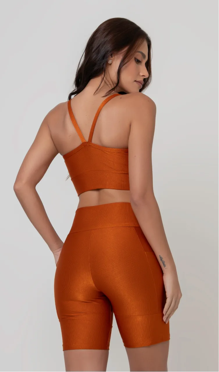 Uma modelo feminina posando de costas contra um fundo cinza neutro, vestindo um conjunto fitness feminino laranja, com proteção solar. O top fitness de academia tem alças finas. A peça de baixo é um short de ciclismo (bermuda fitness feminina) de cintura alta também na cor laranja, com um bolso lateral visível onde a modelo está com a mão direita inserida. Ela olha para o lado. 