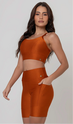 Uma modelo feminina posando de frente contra um fundo cinza neutro, vestindo um conjunto fitness feminino laranja, com proteção solar. O top fitness de academia tem alças finas. A peça de baixo é um short de ciclismo (bermuda fitness feminina) de cintura alta também na cor laranja, com um bolso lateral visível onde a modelo está com a mão direita inserida. Ela olha para o lado. 