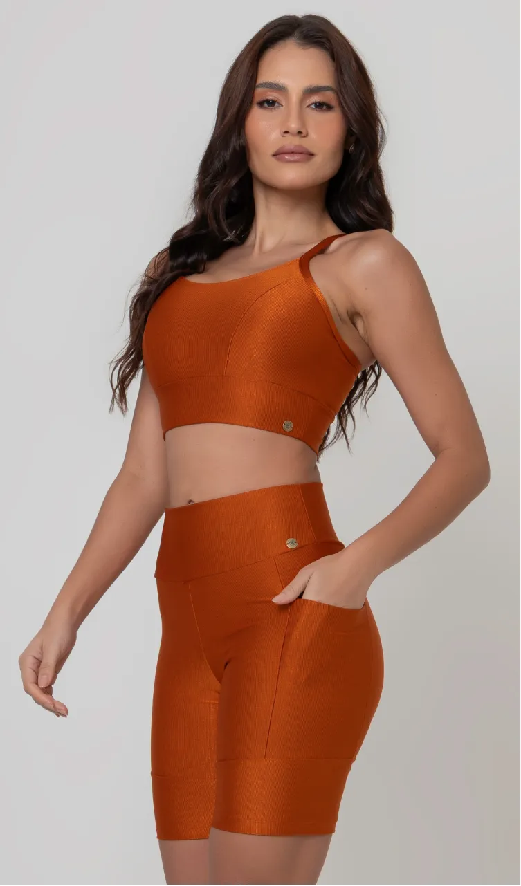 Uma modelo feminina posando de frente contra um fundo cinza neutro, vestindo um conjunto fitness feminino laranja, com proteção solar. O top fitness de academia tem alças finas. A peça de baixo é um short de ciclismo (bermuda fitness feminina) de cintura alta também na cor laranja, com um bolso lateral visível onde a modelo está com a mão direita inserida. Ela olha para o lado. 
