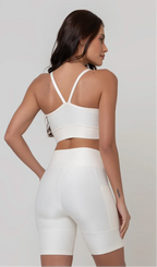 Uma modelo feminina posando de costas contra um fundo cinza neutro, vestindo um conjunto fitness feminino branco, com proteção solar. O top fitness de academia tem alças finas. A peça de baixo é um short de ciclismo (bermuda fitness feminina) de cintura alta também na cor branca, com um bolso lateral visível onde a modelo está com a mão direita inserida. Ela olha para o lado. 
