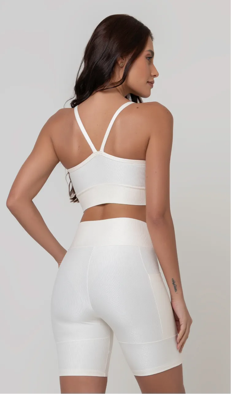 Uma modelo feminina posando de costas contra um fundo cinza neutro, vestindo um conjunto fitness feminino branco, com proteção solar. O top fitness de academia tem alças finas. A peça de baixo é um short de ciclismo (bermuda fitness feminina) de cintura alta também na cor branca, com um bolso lateral visível onde a modelo está com a mão direita inserida. Ela olha para o lado. 