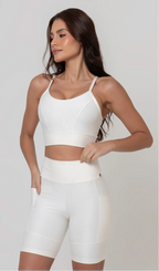 Uma modelo feminina posando de frente contra um fundo cinza neutro, vestindo um conjunto fitness feminino branco, com proteção solar. O top fitness de academia tem alças finas. A peça de baixo é um short de ciclismo (bermuda fitness feminina) de cintura alta também na cor branca, com um bolso lateral visível onde a modelo está com a mão direita inserida. Ela olha para o lado. 