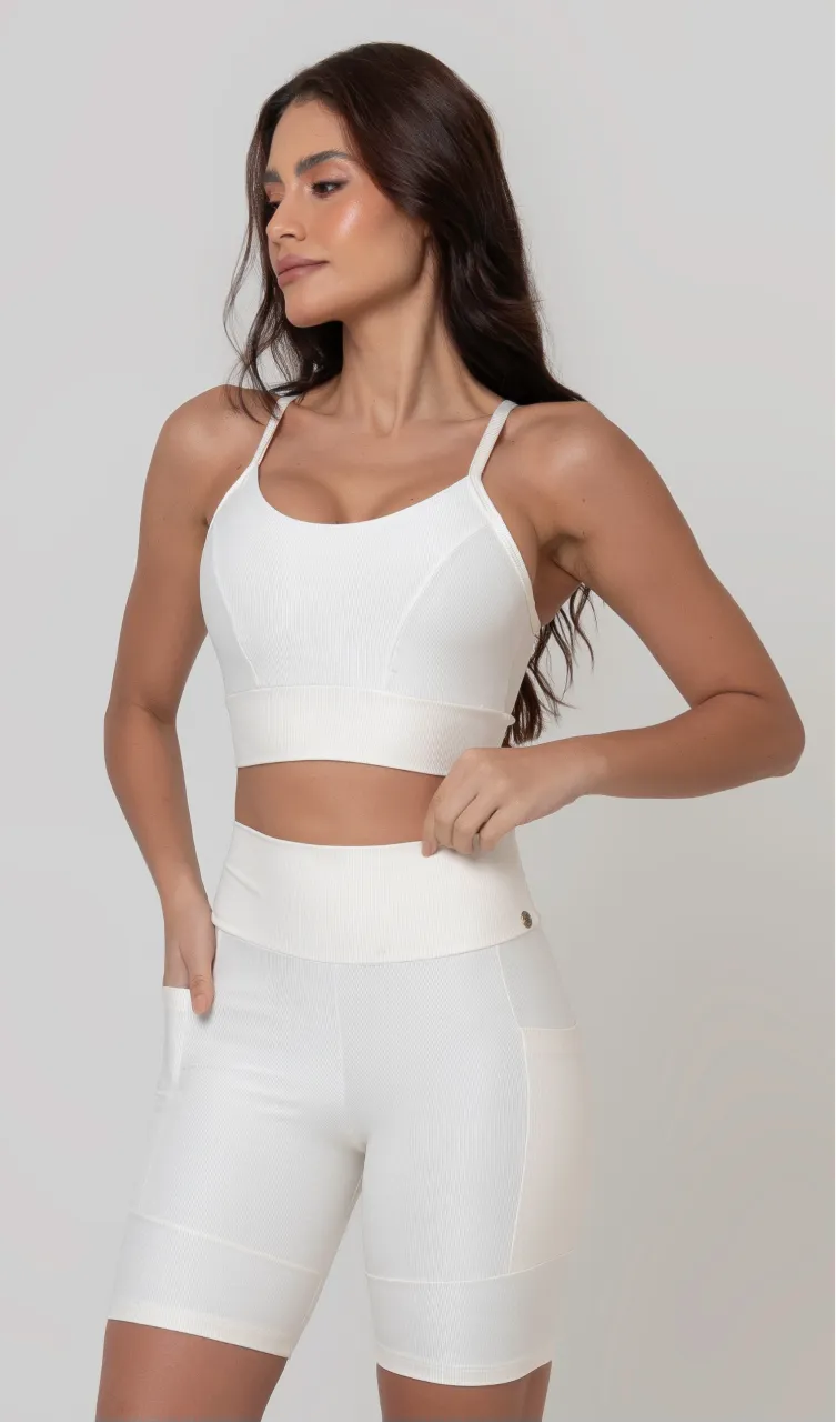 Uma modelo feminina posando de frente contra um fundo cinza neutro, vestindo um conjunto fitness feminino branco, com proteção solar. O top fitness de academia tem alças finas. A peça de baixo é um short de ciclismo (bermuda fitness feminina) de cintura alta também na cor branca, com um bolso lateral visível onde a modelo está com a mão direita inserida. Ela olha para o lado. 