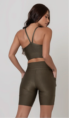Uma modelo feminina posando de costas contra um fundo cinza neutro, vestindo um conjunto fitness feminino verde, com proteção solar. O top fitness de academia tem alças finas. A peça de baixo é um short de ciclismo (bermuda fitness feminina) de cintura alta também na cor verde, com um bolso lateral visível onde a modelo está com a mão direita inserida. Ela olha para o lado. 