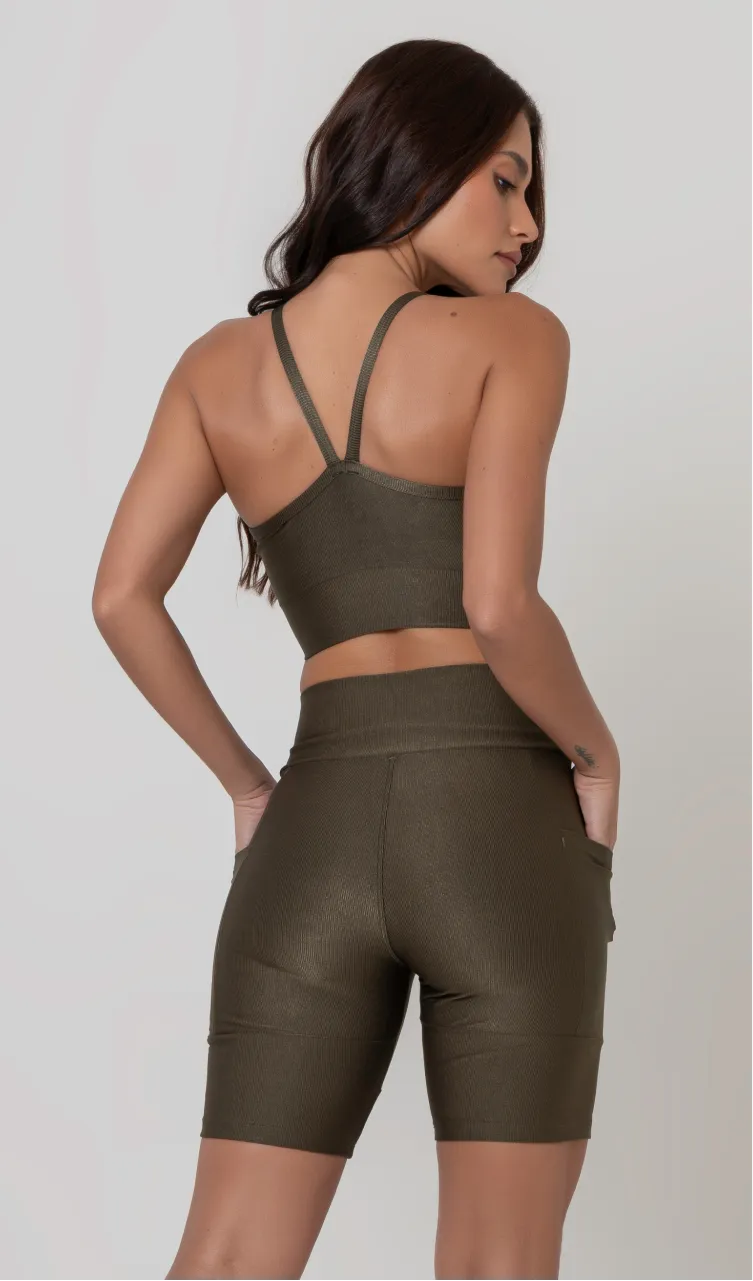 Uma modelo feminina posando de costas contra um fundo cinza neutro, vestindo um conjunto fitness feminino verde, com proteção solar. O top fitness de academia tem alças finas. A peça de baixo é um short de ciclismo (bermuda fitness feminina) de cintura alta também na cor verde, com um bolso lateral visível onde a modelo está com a mão direita inserida. Ela olha para o lado. 