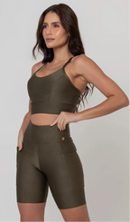 Uma modelo feminina posando de frente contra um fundo cinza neutro, vestindo um conjunto fitness feminino verde, com proteção solar. O top fitness de academia tem alças finas. A peça de baixo é um short de ciclismo (bermuda fitness feminina) de cintura alta também na cor verde, com um bolso lateral visível onde a modelo está com a mão direita inserida. Ela olha para o lado. 