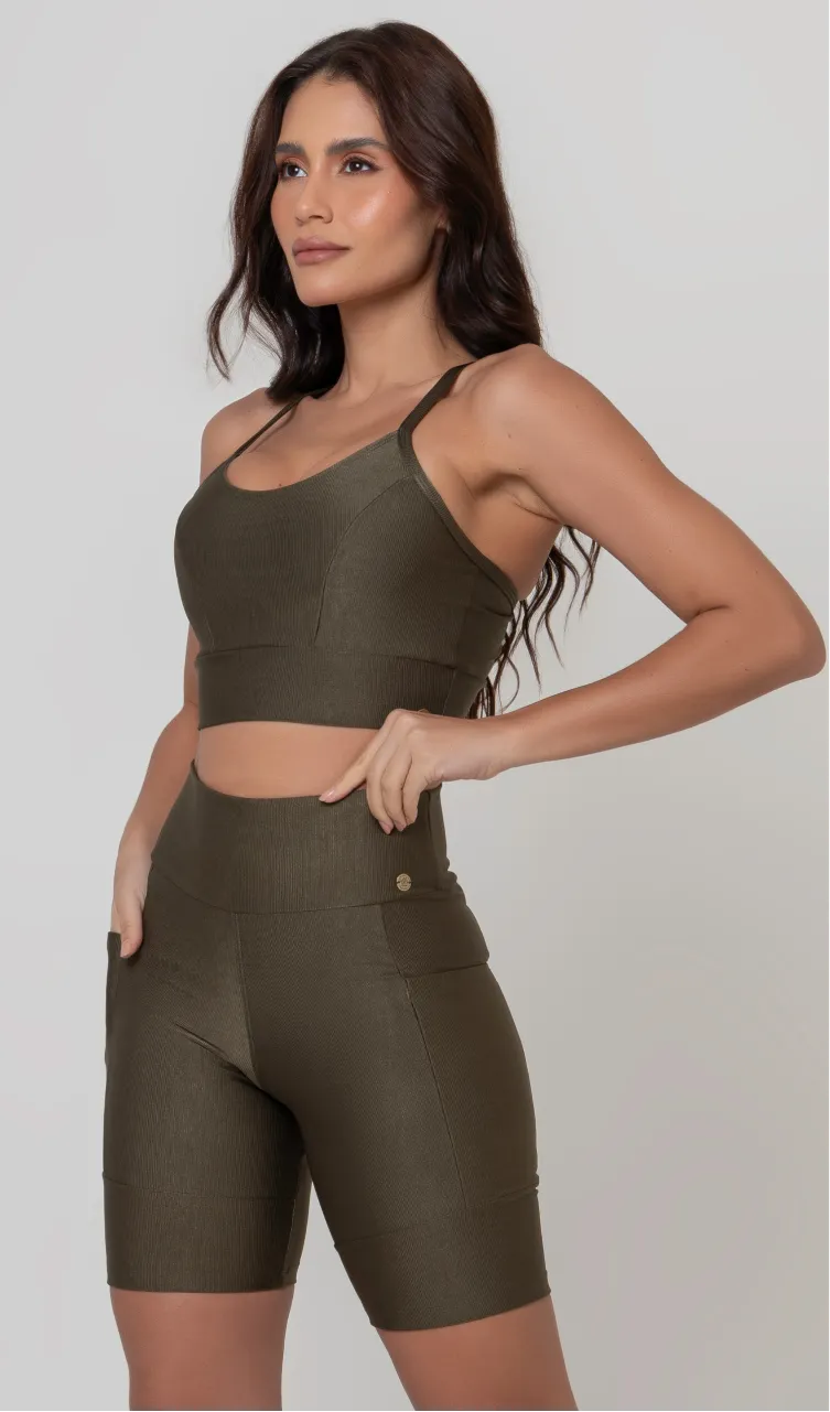 Uma modelo feminina posando de frente contra um fundo cinza neutro, vestindo um conjunto fitness feminino verde, com proteção solar. O top fitness de academia tem alças finas. A peça de baixo é um short de ciclismo (bermuda fitness feminina) de cintura alta também na cor verde, com um bolso lateral visível onde a modelo está com a mão direita inserida. Ela olha para o lado. 