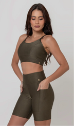 Uma modelo feminina posando de frente contra um fundo cinza neutro, vestindo um conjunto fitness feminino verde, com proteção solar. O top fitness de academia tem alças finas. A peça de baixo é um short de ciclismo (bermuda fitness feminina) de cintura alta também na cor verde, com um bolso lateral visível onde a modelo está com a mão direita inserida. Ela olha para baixo.
