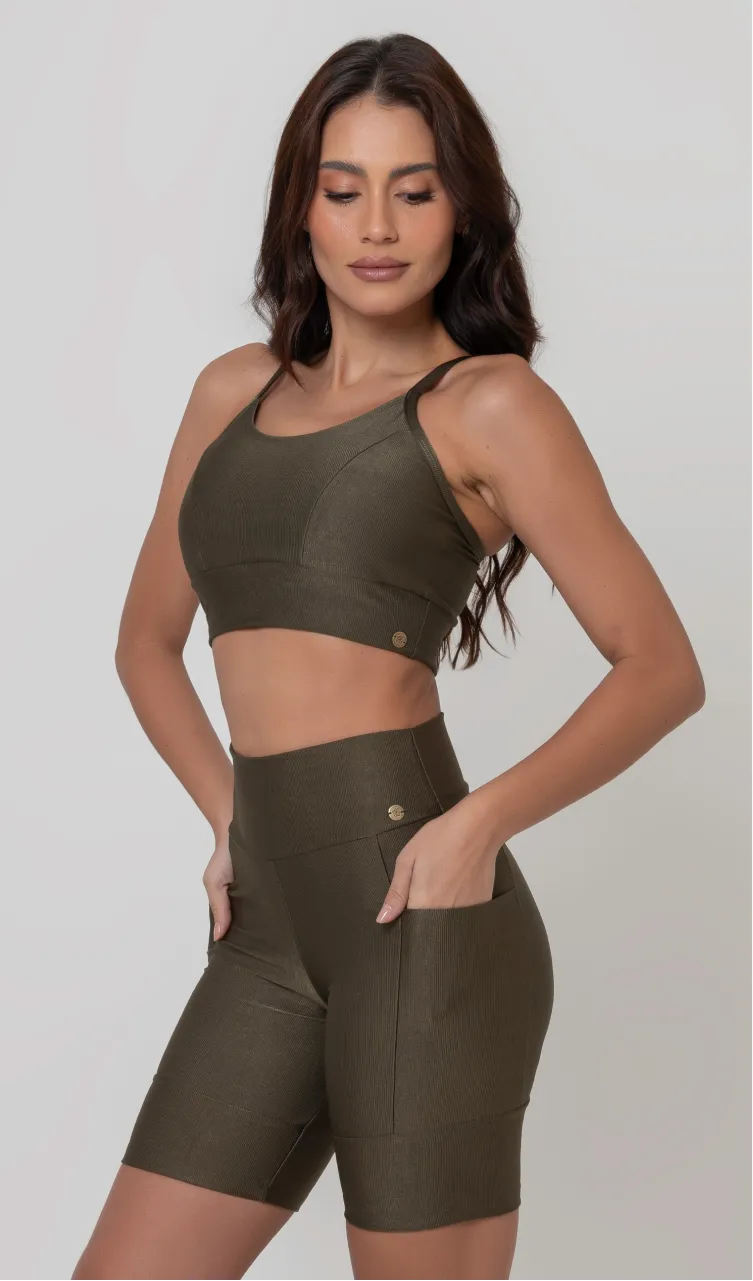 Uma modelo feminina posando de frente contra um fundo cinza neutro, vestindo um conjunto fitness feminino verde, com proteção solar. O top fitness de academia tem alças finas. A peça de baixo é um short de ciclismo (bermuda fitness feminina) de cintura alta também na cor verde, com um bolso lateral visível onde a modelo está com a mão direita inserida. Ela olha para baixo.