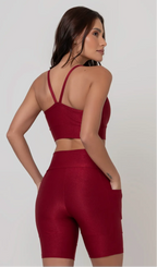 Uma modelo feminina posando de costas contra um fundo cinza neutro, vestindo um conjunto fitness feminino vermelho, com proteção solar. O top fitness de academia tem alças finas. A peça de baixo é um short de ciclismo (bermuda fitness feminina) de cintura alta também na cor vermelha, com um bolso lateral visível onde a modelo está com a mão direita inserida. Ela olha para o lado. 