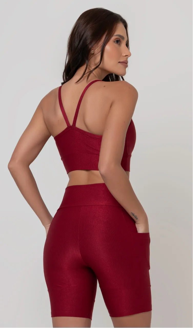 Uma modelo feminina posando de costas contra um fundo cinza neutro, vestindo um conjunto fitness feminino vermelho, com proteção solar. O top fitness de academia tem alças finas. A peça de baixo é um short de ciclismo (bermuda fitness feminina) de cintura alta também na cor vermelha, com um bolso lateral visível onde a modelo está com a mão direita inserida. Ela olha para o lado. 