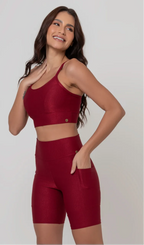 Uma modelo feminina posando de frente contra um fundo cinza neutro, vestindo um conjunto fitness feminino vermelho, com proteção solar. O top fitness de academia tem alças finas. A peça de baixo é um short de ciclismo (bermuda fitness feminina) de cintura alta também na cor vermelha, com um bolso lateral visível onde a modelo está com a mão direita inserida. Ela olha para o lado. 