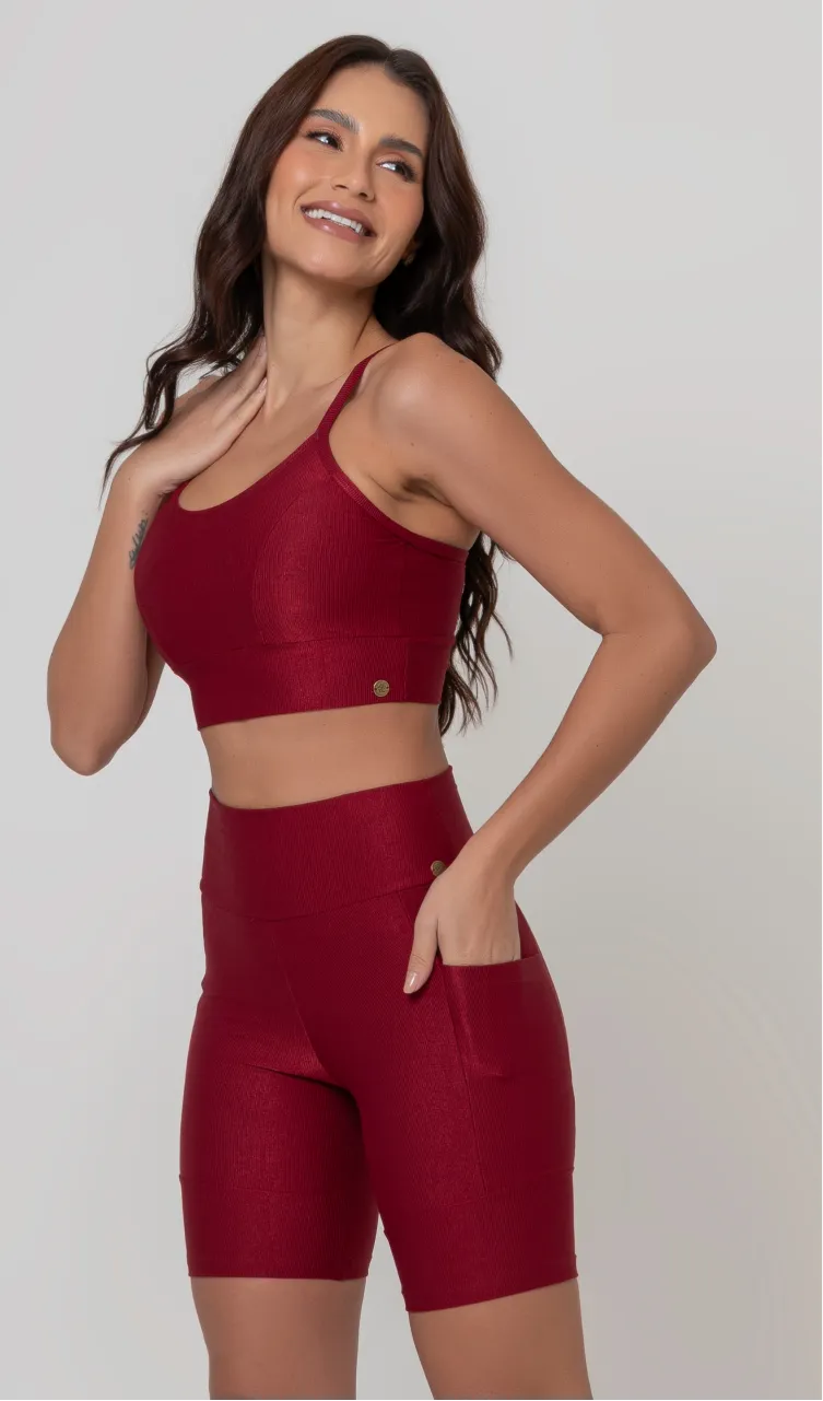 Uma modelo feminina posando de frente contra um fundo cinza neutro, vestindo um conjunto fitness feminino vermelho, com proteção solar. O top fitness de academia tem alças finas. A peça de baixo é um short de ciclismo (bermuda fitness feminina) de cintura alta também na cor vermelha, com um bolso lateral visível onde a modelo está com a mão direita inserida. Ela olha para o lado. 