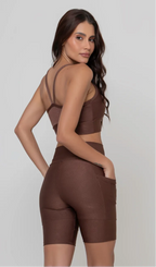 Uma modelo feminina posando de costas contra um fundo cinza neutro, vestindo um conjunto fitness feminino marrom, com proteção solar. O top fitness de academia tem alças finas. A peça de baixo é um short de ciclismo (bermuda fitness feminina) de cintura alta também na cor marrom, com um bolso lateral visível onde a modelo está com a mão direita inserida. Ela olha para o lado. 