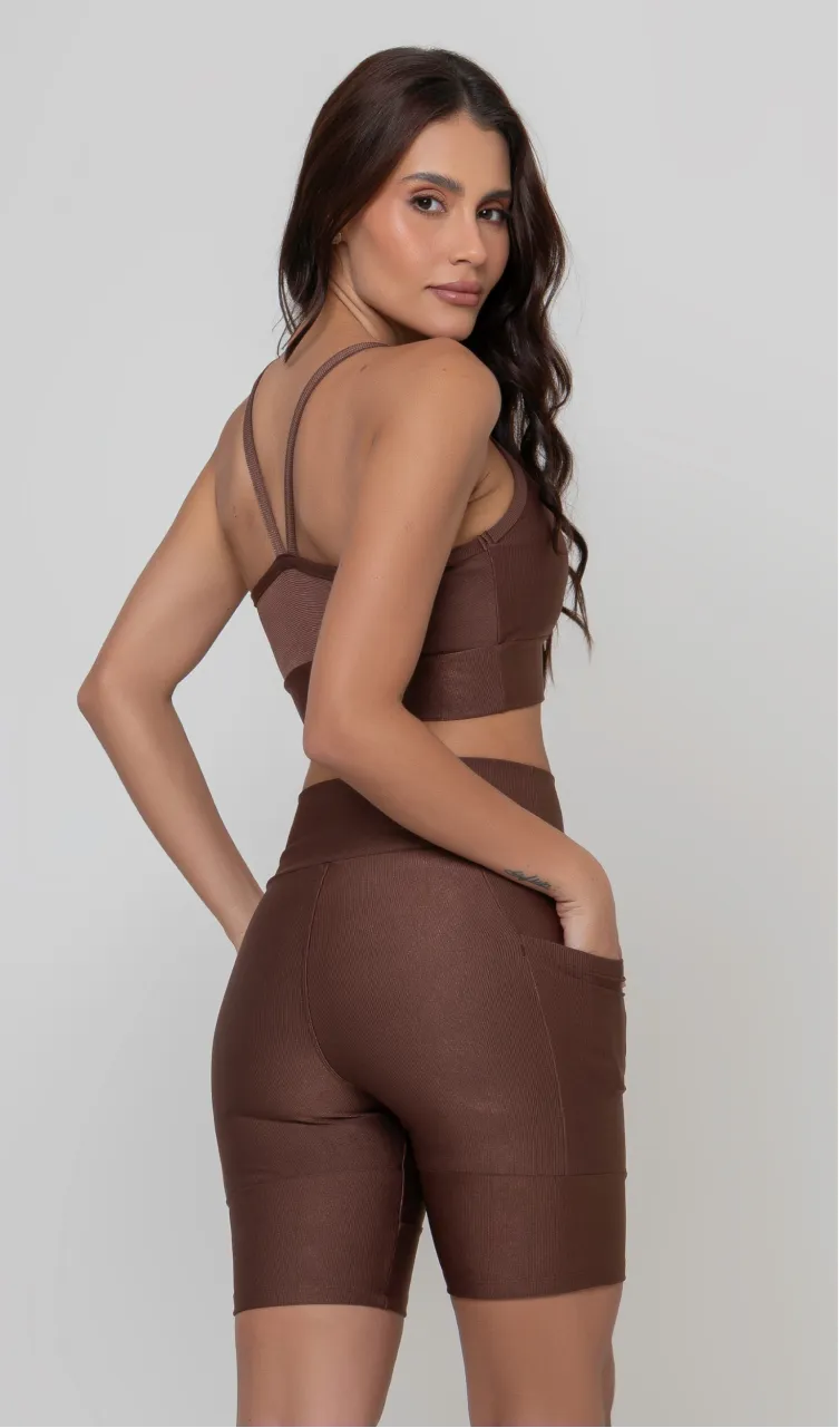 Uma modelo feminina posando de costas contra um fundo cinza neutro, vestindo um conjunto fitness feminino marrom, com proteção solar. O top fitness de academia tem alças finas. A peça de baixo é um short de ciclismo (bermuda fitness feminina) de cintura alta também na cor marrom, com um bolso lateral visível onde a modelo está com a mão direita inserida. Ela olha para o lado. 