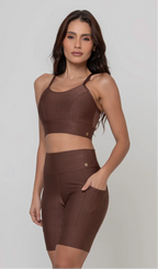 Uma modelo feminina posando de frente contra um fundo cinza neutro, vestindo um conjunto fitness feminino marrom, com proteção solar. O top fitness de academia tem alças finas. A peça de baixo é um short de ciclismo (bermuda fitness feminina) de cintura alta também na cor marrom, com um bolso lateral visível onde a modelo está com a mão direita inserida. Ela olha para o lado. 