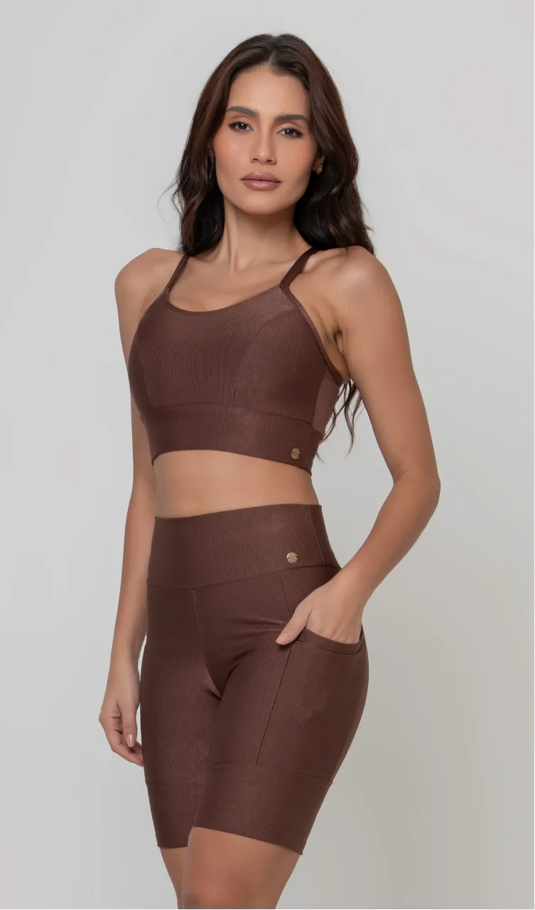 Uma modelo feminina posando de frente contra um fundo cinza neutro, vestindo um conjunto fitness feminino marrom, com proteção solar. O top fitness de academia tem alças finas. A peça de baixo é um short de ciclismo (bermuda fitness feminina) de cintura alta também na cor marrom, com um bolso lateral visível onde a modelo está com a mão direita inserida. Ela olha para o lado. 