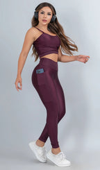 Modelo feminina usando conjunto fitness feminino roxo uva Mi Raromi de duas peças. Inclui um top cropped de alcinhas finas e leggings de cintura alta correspondentes, ambos feitos de poliamida e elastano. A modelo está em pé contra um fundo cinza neutro, usando tênis brancos.