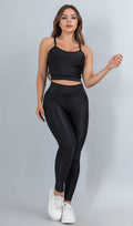 Modelo feminina usando conjunto fitness feminino preto Mi Raromi de duas peças. Inclui um top cropped de alcinhas finas e leggings de cintura alta correspondentes, ambos feitos de poliamida e elastano. A modelo está em pé contra um fundo cinza neutro, usando tênis brancos.
