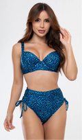 Uma mulher de frente, com cabelos escuros vestindo um conjunto de biquíni de duas peças. O biquíni tem uma estampa colorida com tema animal print preto em fundo azul. Top estruturado com aro e bojo, sendo um sutiã meia taça. A calcinha é de cintura alta hot pant com cordões laterais para amarração detalhados. O fundo é cinza neutro. 