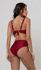 Uma mulher de costas, com cabelos escuros vestindo um  biquíni vermelho. Top estruturado com aro e bojo, sendo um sutiã meia taça. A calcinha é de cintura alta hot pant com cordões laterais para amarração detalhados. O fundo é cinza neutro. 