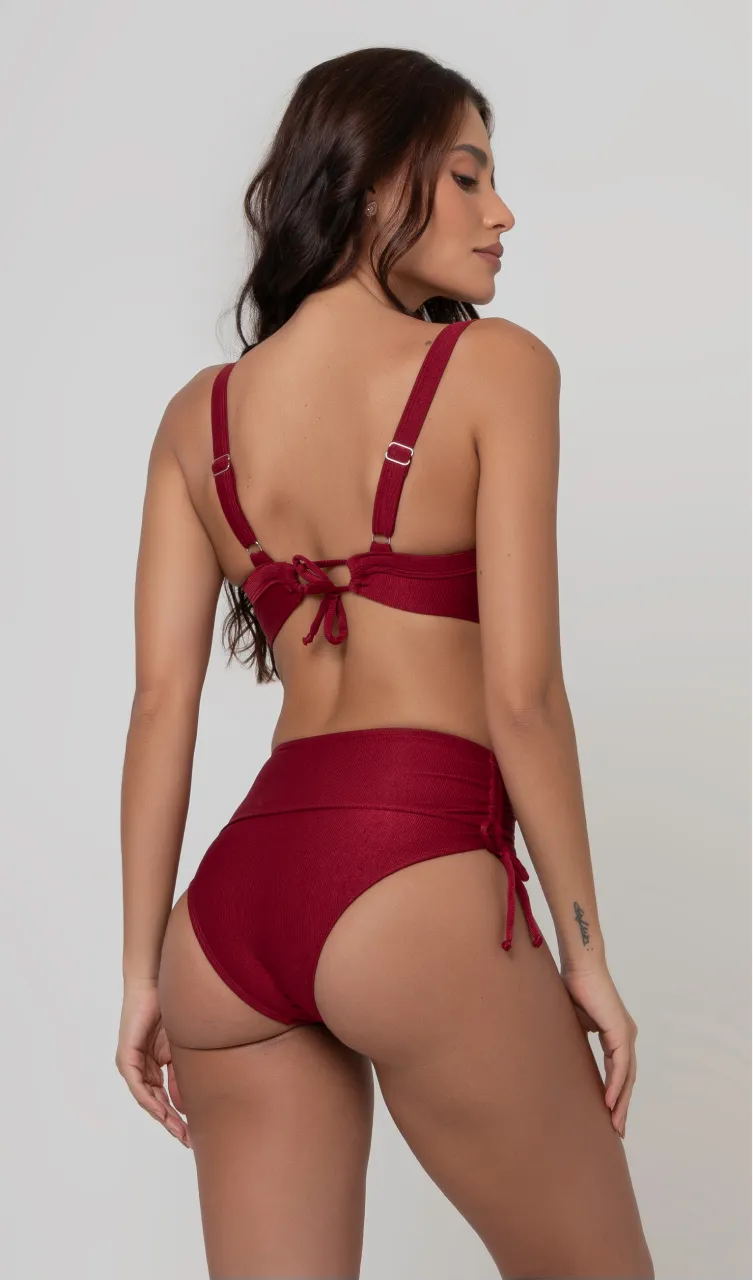 Uma mulher de costas, com cabelos escuros vestindo um  biquíni vermelho. Top estruturado com aro e bojo, sendo um sutiã meia taça. A calcinha é de cintura alta hot pant com cordões laterais para amarração detalhados. O fundo é cinza neutro. 