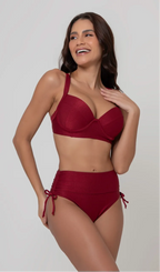 Uma mulher de frente, com cabelos escuros vestindo um  biquíni vermelho. Top estruturado com aro e bojo, sendo um sutiã meia taça. A calcinha é de cintura alta hot pant com cordões laterais para amarração detalhados. O fundo é cinza neutro. 