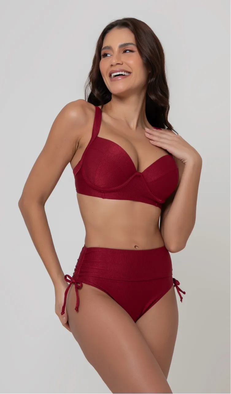 Uma mulher de frente, com cabelos escuros vestindo um  biquíni vermelho. Top estruturado com aro e bojo, sendo um sutiã meia taça. A calcinha é de cintura alta hot pant com cordões laterais para amarração detalhados. O fundo é cinza neutro. 