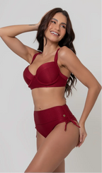 Uma mulher de frente, com cabelos escuros vestindo um  biquíni vermelho. Top estruturado com aro e bojo, sendo um sutiã meia taça. A calcinha é de cintura alta hot pant com cordões laterais para amarração detalhados. O fundo é cinza neutro. 