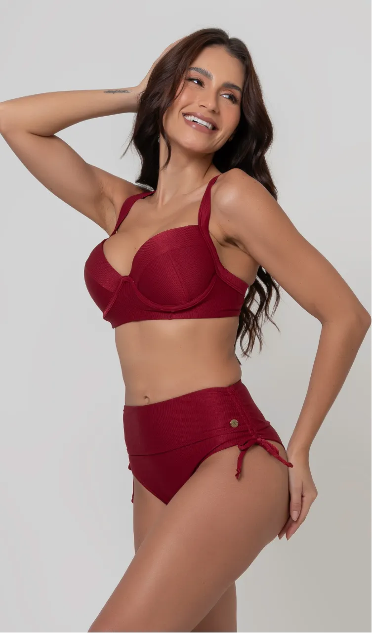 Uma mulher de frente, com cabelos escuros vestindo um  biquíni vermelho. Top estruturado com aro e bojo, sendo um sutiã meia taça. A calcinha é de cintura alta hot pant com cordões laterais para amarração detalhados. O fundo é cinza neutro. 