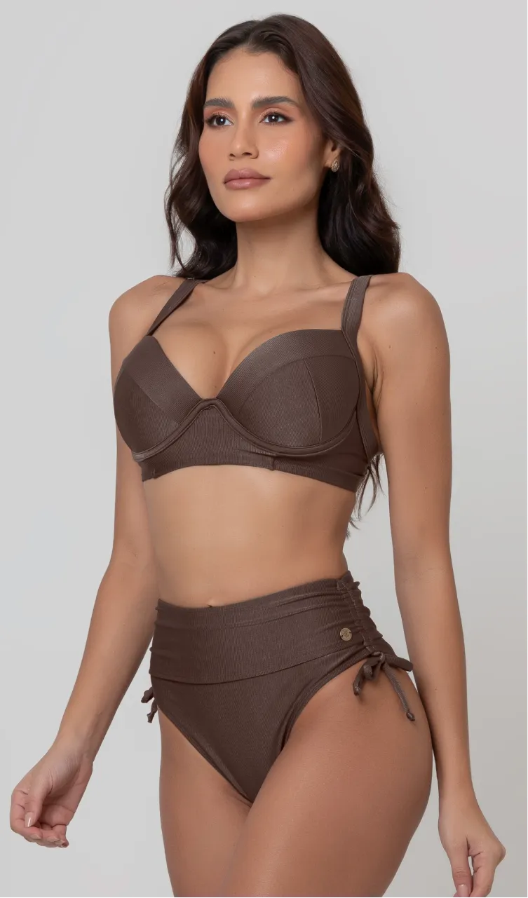 Uma mulher de frente, com cabelos escuros vestindo um biquíni marrom. Top estruturado com aro e bojo, sendo um sutiã meia taça. A calcinha é de cintura alta hot pant com cordões laterais para amarração detalhados. O fundo é cinza neutro. 
