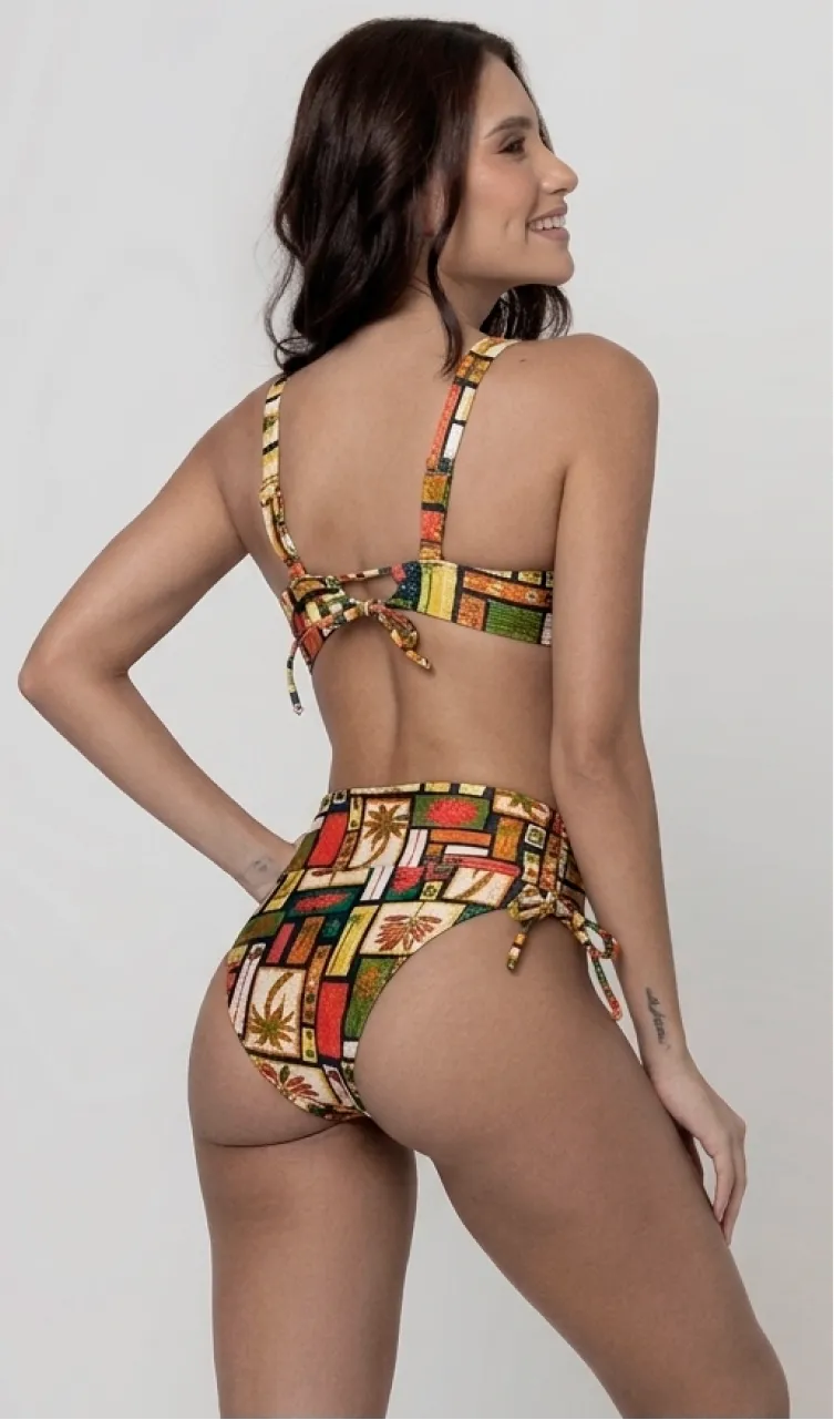 Uma mulher de costas, com cabelos escuros vestindo um conjunto de biquíni de duas peças. O biquíni tem uma estampa colorida com temas tropicais e ícones. Top estruturado com aro e bojo, sendo um sutiã meia taça. A calcinha é de cintura alta hot pant com cordões laterais para amarração detalhados. O fundo é cinza neutro. 