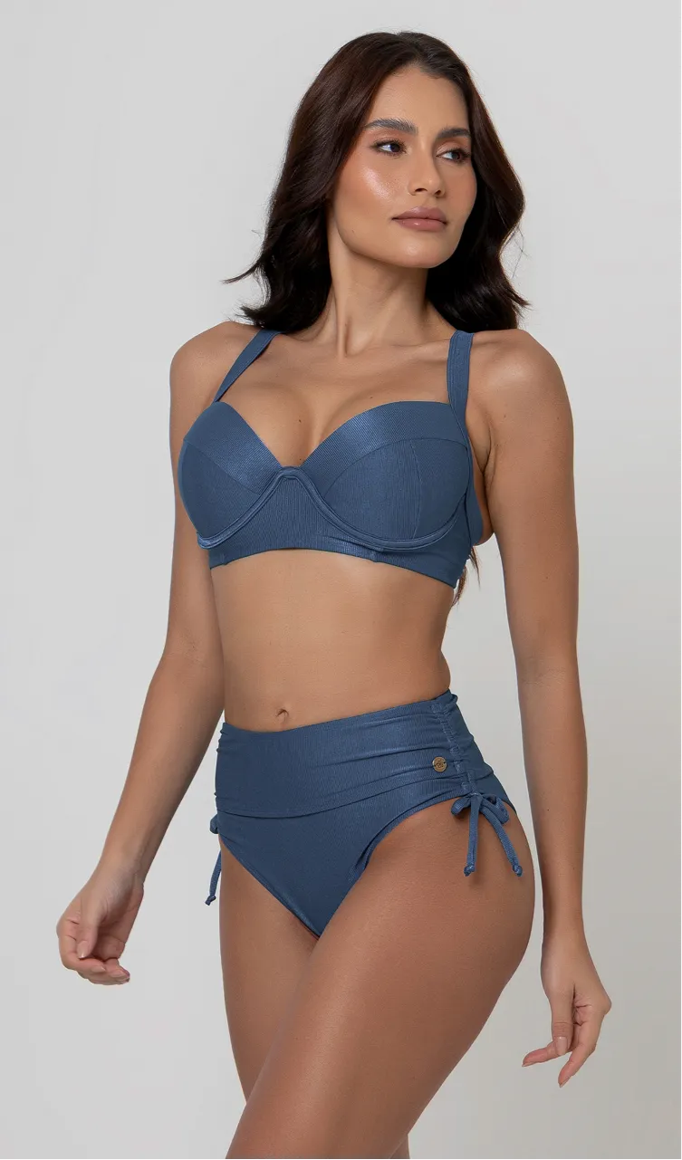 Uma mulher de frente, com cabelos escuros vestindo um biquíni safira. Top estruturado com aro e bojo, sendo um sutiã meia taça. A calcinha é de cintura alta hot pant com cordões laterais para amarração detalhados. O fundo é cinza neutro. 