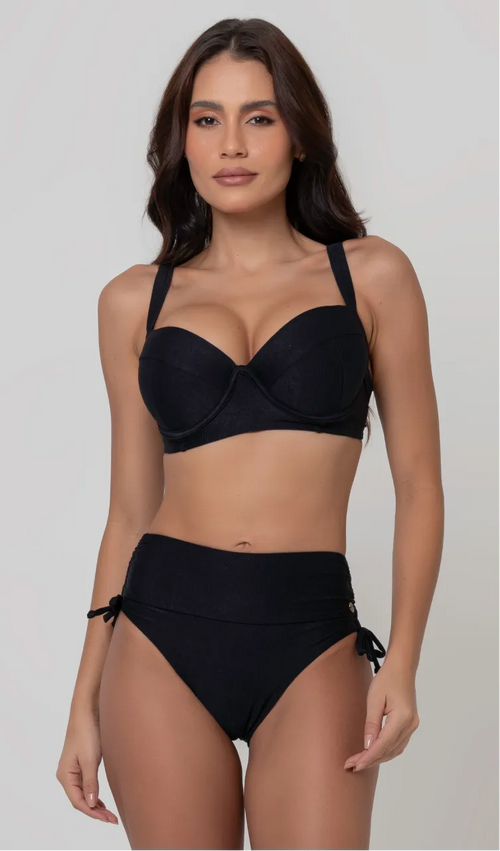Uma mulher de frente, com cabelos escuros vestindo um  biquíni preto. Top estruturado com aro e bojo, sendo um sutiã meia taça. A calcinha é de cintura alta hot pant com cordões laterais para amarração detalhados. O fundo é cinza neutro. 