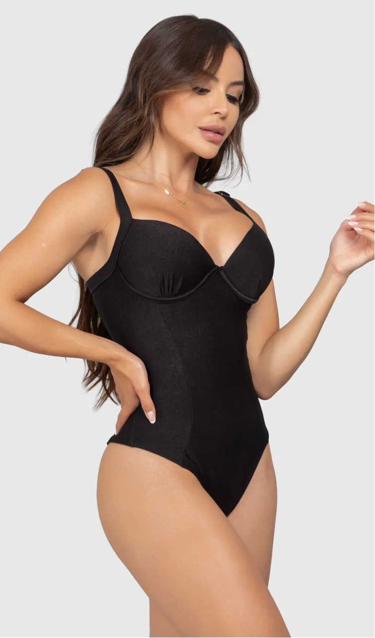 Modelo feminina de cabelos claros, de lado, em um estúdio com fundo neutro, vestindo um maiô preto com efeito modelador. A peça é confeccionada em tecido de poliamida e elastano, com proteção solar. Maiô estruturado com sutiã meia taça com aro para sustentação, bojo, decote coração e amarração nas costas estilo espartilho.