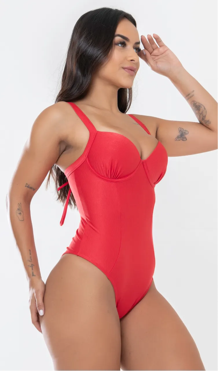 Modelo feminina de cabelos claros, de lado, em um estúdio com fundo neutro, vestindo um maiô vermelho melancia com efeito modelador. A peça é confeccionada em tecido de poliamida e elastano, com proteção solar. Maiô estruturado com sutiã meia taça com aro para sustentação, bojo, decote coração e amarração nas costas estilo espartilho.