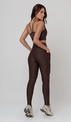 Uma foto de estúdio de corpo inteiro de uma mulher jovem posando de costas com um conjunto fitness feminino feito de poliamida e elastano na cor marrom. O conjunto é composto por um top de alças finas, bojo removível e uma calça legging de cintura alta com bolsos laterais práticos. Ela está com uma mão no bolso da legging e usa tênis esportivos cinzas e bege. O fundo é cinza claro e bem iluminado. Este conjunto de treino preto é ideal para academia e exercícios físicos.