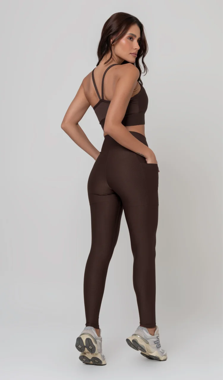 Uma foto de estúdio de corpo inteiro de uma mulher jovem posando de costas com um conjunto fitness feminino feito de poliamida e elastano na cor marrom. O conjunto é composto por um top de alças finas, bojo removível e uma calça legging de cintura alta com bolsos laterais práticos. Ela está com uma mão no bolso da legging e usa tênis esportivos cinzas e bege. O fundo é cinza claro e bem iluminado. Este conjunto de treino preto é ideal para academia e exercícios físicos.