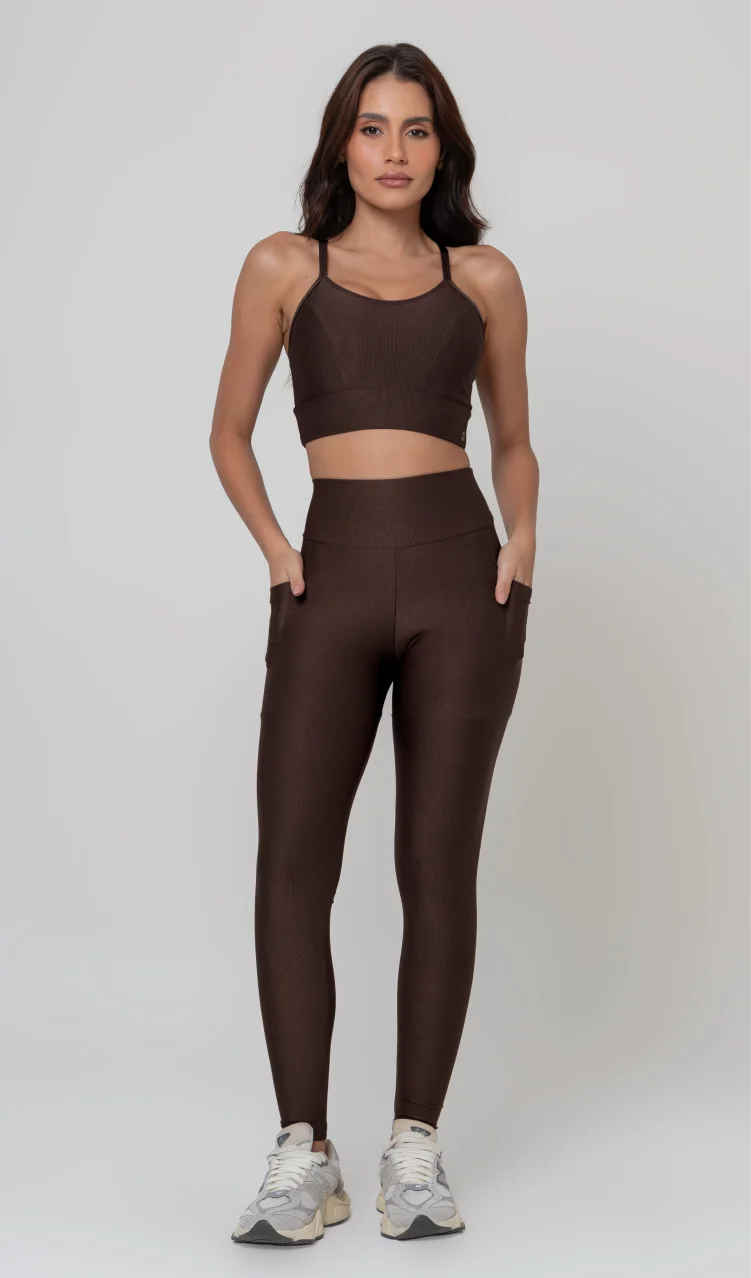 Uma foto de estúdio de corpo inteiro de uma mulher jovem posando com um conjunto fitness feminino feito de poliamida e elastano na cor marrom. O conjunto é composto por um top de alças finas, bojo removível e uma calça legging de cintura alta com bolsos laterais práticos. Ela está com uma mão no bolso da legging e usa tênis esportivos cinzas e bege. O fundo é cinza claro e bem iluminado. Este conjunto de treino preto é ideal para academia e exercícios físicos.