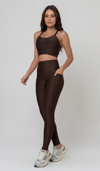 Uma foto de estúdio de corpo inteiro de uma mulher jovem posando com um conjunto fitness feminino feito de poliamida e elastano na cor marrom. O conjunto é composto por um top de alças finas, bojo removível e uma calça legging de cintura alta com bolsos laterais práticos. Ela está com uma mão no bolso da legging e usa tênis esportivos cinzas e bege. O fundo é cinza claro e bem iluminado. Este conjunto de treino preto é ideal para academia e exercícios físicos.