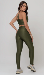Uma foto de estúdio de corpo inteiro de uma mulher jovem posando de costas com um conjunto fitness feminino feito de poliamida e elastano na cor verde. O conjunto é composto por um top de alças finas, bojo removível e uma calça legging de cintura alta com bolsos laterais práticos. Ela está com uma mão no bolso da legging e usa tênis esportivos cinzas e bege. O fundo é cinza claro e bem iluminado. Este conjunto de treino preto é ideal para academia e exercícios físicos.