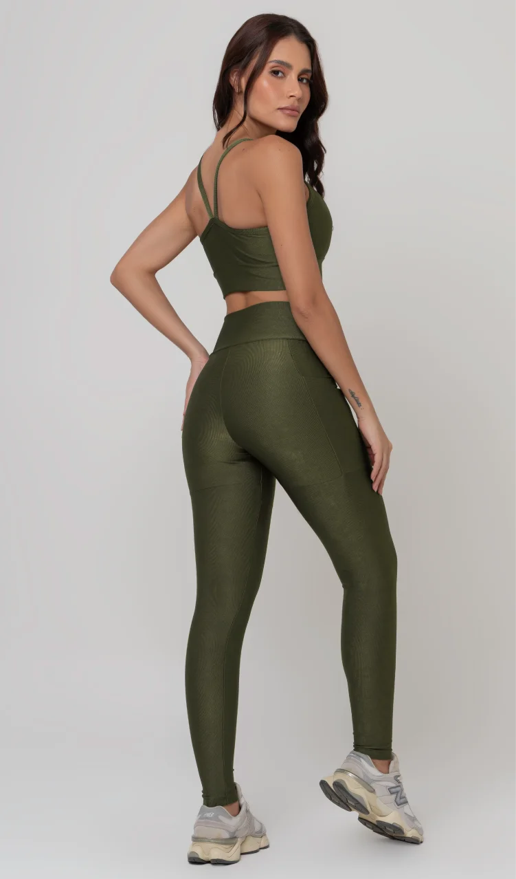 Uma foto de estúdio de corpo inteiro de uma mulher jovem posando de costas com um conjunto fitness feminino feito de poliamida e elastano na cor verde. O conjunto é composto por um top de alças finas, bojo removível e uma calça legging de cintura alta com bolsos laterais práticos. Ela está com uma mão no bolso da legging e usa tênis esportivos cinzas e bege. O fundo é cinza claro e bem iluminado. Este conjunto de treino preto é ideal para academia e exercícios físicos.