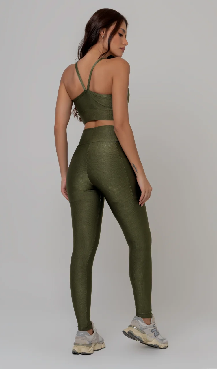 Uma foto de estúdio de corpo inteiro de uma mulher jovem posando de costas com um conjunto fitness feminino feito de poliamida e elastano na cor verde. O conjunto é composto por um top de alças finas, bojo removível e uma calça legging de cintura alta com bolsos laterais práticos. Ela está com uma mão no bolso da legging e usa tênis esportivos cinzas e bege. O fundo é cinza claro e bem iluminado. Este conjunto de treino preto é ideal para academia e exercícios físicos.