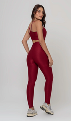Uma foto de estúdio de corpo inteiro de uma mulher jovem posando de costas com um conjunto fitness feminino feito de poliamida e elastano na cor vermelha. O conjunto é composto por um top de alças finas, bojo removível e uma calça legging de cintura alta com bolsos laterais práticos. Ela está com uma mão no bolso da legging e usa tênis esportivos cinzas e bege. O fundo é cinza claro e bem iluminado. Este conjunto de treino preto é ideal para academia e exercícios físicos.
