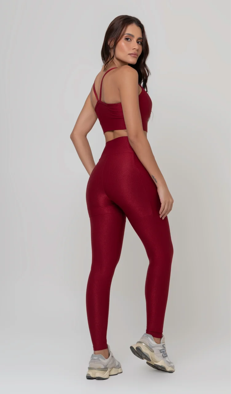 Uma foto de estúdio de corpo inteiro de uma mulher jovem posando de costas com um conjunto fitness feminino feito de poliamida e elastano na cor vermelha. O conjunto é composto por um top de alças finas, bojo removível e uma calça legging de cintura alta com bolsos laterais práticos. Ela está com uma mão no bolso da legging e usa tênis esportivos cinzas e bege. O fundo é cinza claro e bem iluminado. Este conjunto de treino preto é ideal para academia e exercícios físicos.