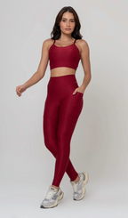Uma foto de estúdio de corpo inteiro de uma mulher jovem posando com um conjunto fitness feminino feito de poliamida e elastano na cor vermelha. O conjunto é composto por um top de alças finas, bojo removível e uma calça legging de cintura alta com bolsos laterais práticos. Ela está com uma mão no bolso da legging e usa tênis esportivos cinzas e bege. O fundo é cinza claro e bem iluminado. Este conjunto de treino preto é ideal para academia e exercícios físicos.
