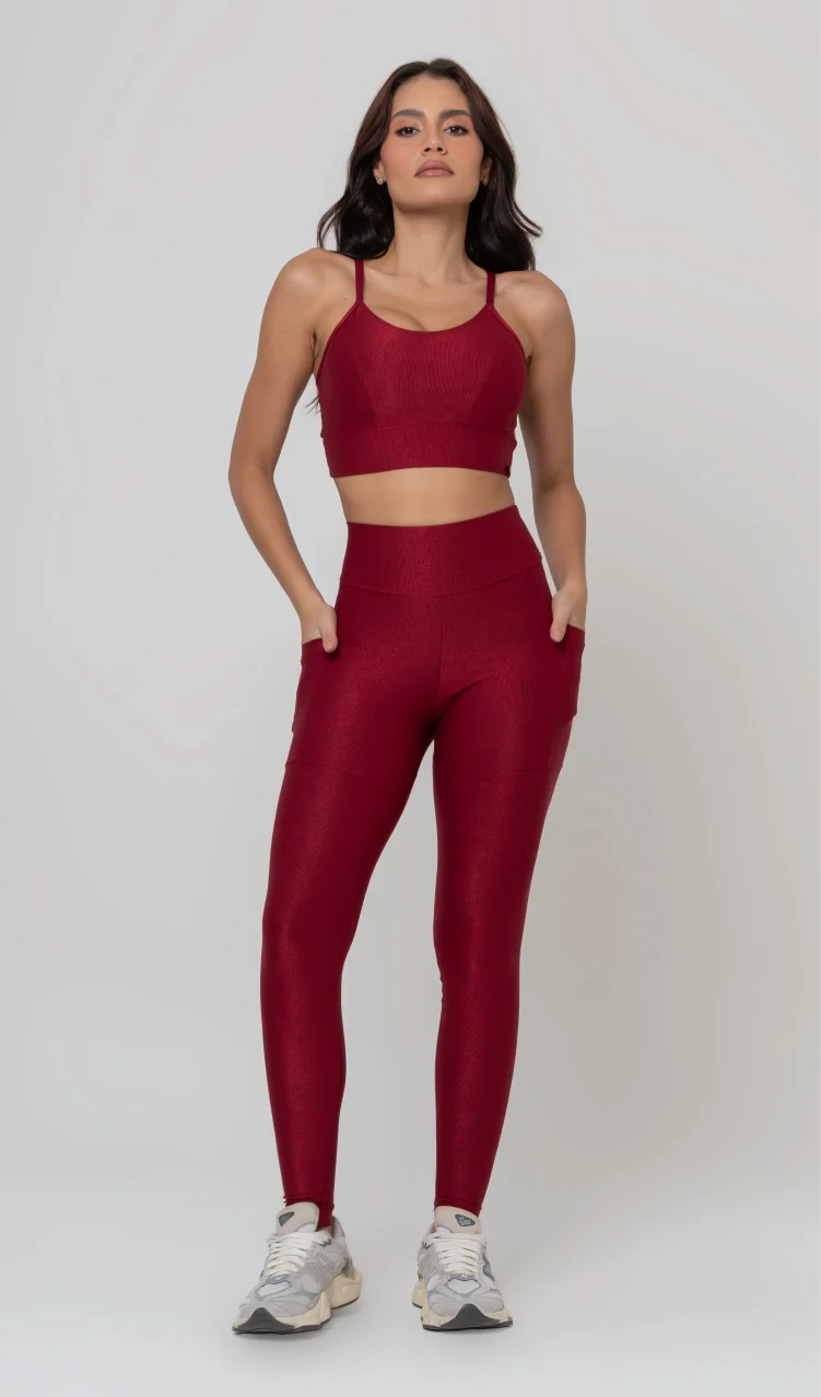 Uma foto de estúdio de corpo inteiro de uma mulher jovem posando com um conjunto fitness feminino feito de poliamida e elastano na cor vermelha. O conjunto é composto por um top de alças finas, bojo removível e uma calça legging de cintura alta com bolsos laterais práticos. Ela está com uma mão no bolso da legging e usa tênis esportivos cinzas e bege. O fundo é cinza claro e bem iluminado. Este conjunto de treino preto é ideal para academia e exercícios físicos.