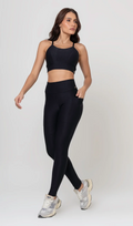 Uma foto de estúdio de corpo inteiro de uma mulher jovem posando com um conjunto fitness feminino feito de poliamida e elastano na cor preta. O conjunto é composto por um top de alças finas, bojo removível e uma calça legging de cintura alta com bolsos laterais práticos. Ela está com uma mão no bolso da legging e usa tênis esportivos cinzas e bege. O fundo é cinza claro e bem iluminado. Este conjunto de treino preto é ideal para academia e exercícios físicos.1