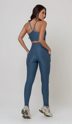 Uma foto de estúdio de corpo inteiro de uma mulher jovem posando com um conjunto fitness feminino feito de poliamida e elastano na cor safira, um tom de azul profundo, fechado e brilhante. O conjunto é composto por um top de alças finas, bojo removível e uma calça legging de cintura alta com bolsos laterais práticos. Ela está com uma mão no bolso da legging e usa tênis esportivos cinzas e bege. O fundo é cinza claro e bem iluminado. Este conjunto de treino preto é ideal para academia e exercícios físicos.